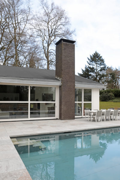 Moderne villa met grote raampartijen, gemetselde schoorsteen en terras aan een zwembad.