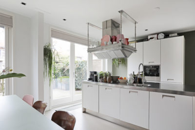 Moderne witte keuken met roestvrijstalen werkblad en hangende afzuigkap in een lichte ruimte met tuinzicht.