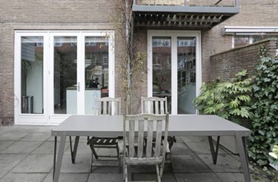 Strakke grijze buitentafel op een terras voor openslaande deuren naar een moderne keuken.