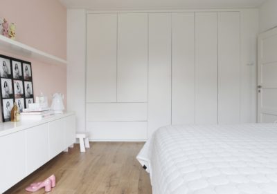 Slaapkamer met maatwerk witte kastenwand, lichte houten vloer en een zachtroze accentmuur.