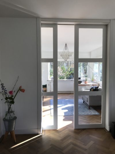 Witte glazen schuifdeuren bieden zicht op een lichte woonkameruitbreiding met panoramisch uitzicht op de tuin.