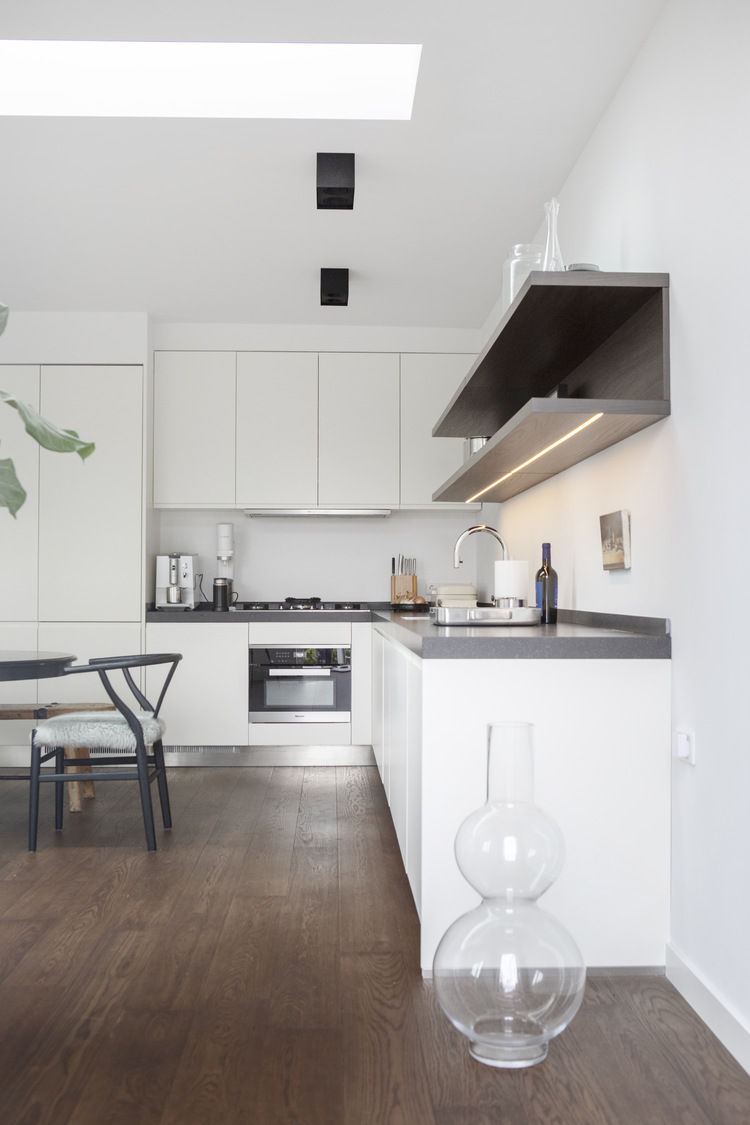 Strakke witte maatwerkkeuken met donker werkblad en houten wandplanken in een vernieuwde woonruimte.