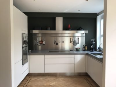 Moderne witte hoekkeuken met RVS achterwand en kookeiland op een klassieke visgraatvloer.